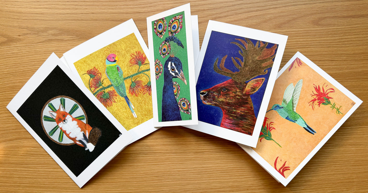 Five colorful art cards with white borders fanned out on a light brown wooden surface. The cards feature various vibrant illustrations including a green bird on a yellow background, a peacock, a deer, and a hummingbird.
